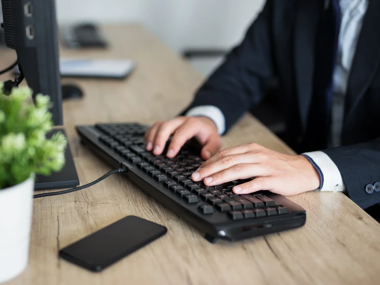 Person im Anzug sitzt am Schreibtisch und tippt etwas in ihren Computer