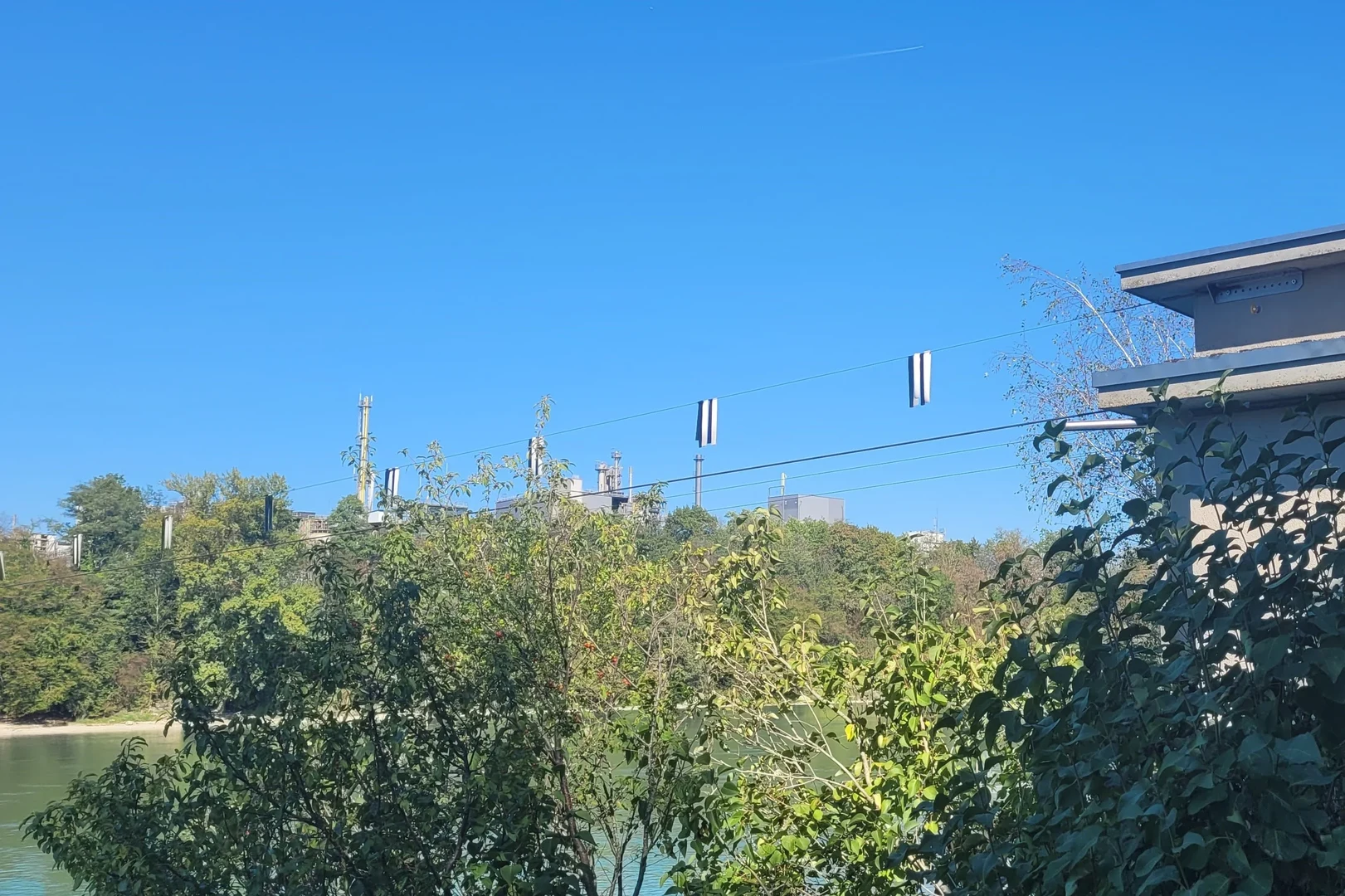 Auf Bild ist ein Gebäude neben einem Fluss zu sehen. Vom Gebäude aus werden mehrere Seil über den Fluss gespannt, an denen zum Teil schwarz-weiße Flatterbänder hängen.  Vor dem Gebäude sind am Ufer im Bild Vordergrund mehrere Büsche zu sehen.