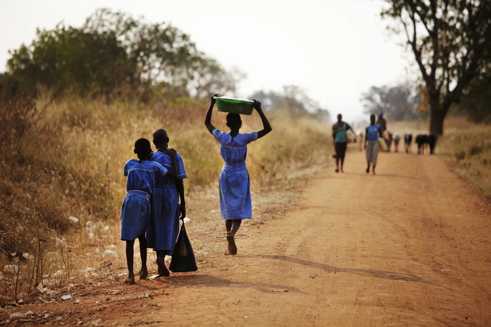 People of Colour Kinder von Hinten auf einer unbefestigten Straße in einem afrikanischen Land, die teilweise Wasserkanister auf ihren Schultern tragen